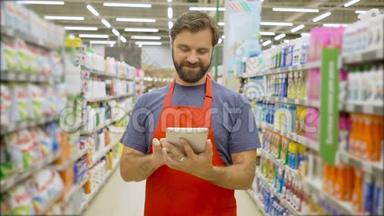 帅气笑脸大胡子超市员工用数码平板电脑站在超市货架上